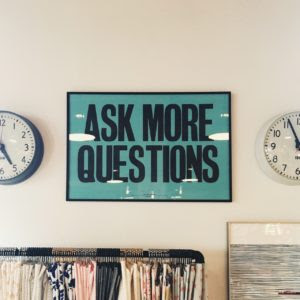 Ask More Questions sign hanging on a wall between two clocks